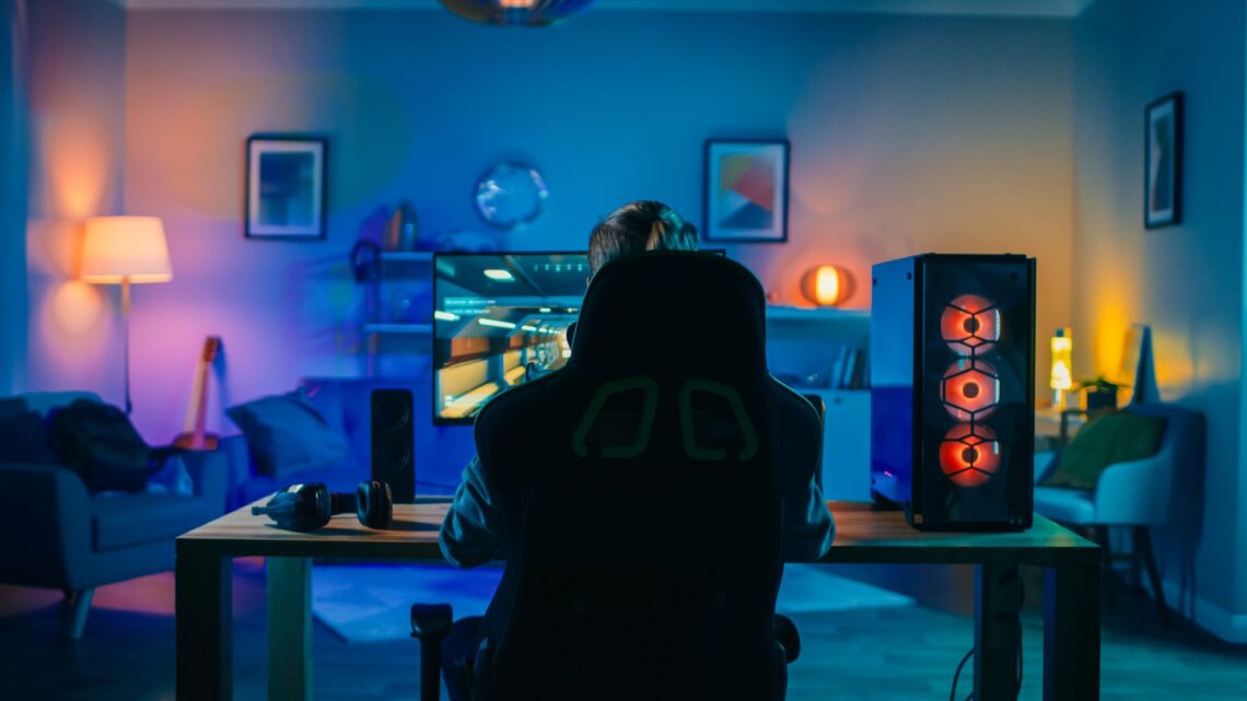 Rückenaufnahme eines Spielers, der ein First-Person-Shooter-Online-Videospiel auf seinem leistungsstarken Computer spielt. Zimmer und PC haben kalte bunte Neon-LED-Lichter. Gemütlicher Abend zu Hause.